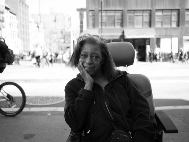 A black and white photograph of a woman smiling sitting in her mobility chair on 14th street.