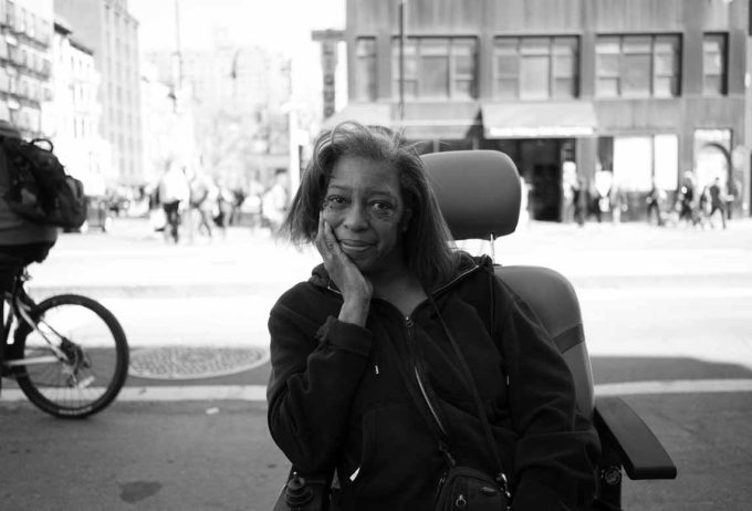 A black and white photograph of a woman smiling sitting in her mobility chair on 14th street.