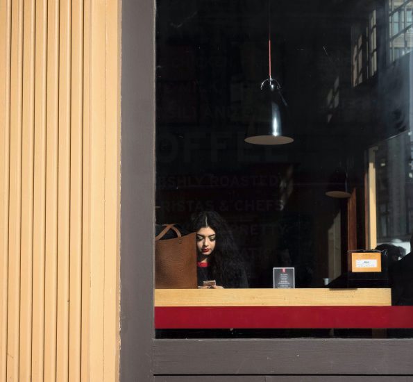 A color photograph of a woman sitting in a coffee shop window wearing lipstick.