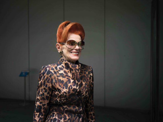 A smiling, glamorous woman with a vivid orange-red bouffant hairstyle is wearing large, dark sunglasses and a high-collared, brown and black leopard print jacket.