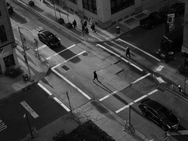 A black and white photograph of people crossing the street with long shadows.