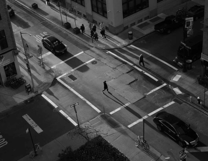 A black and white photograph of people crossing the street with long shadows.
