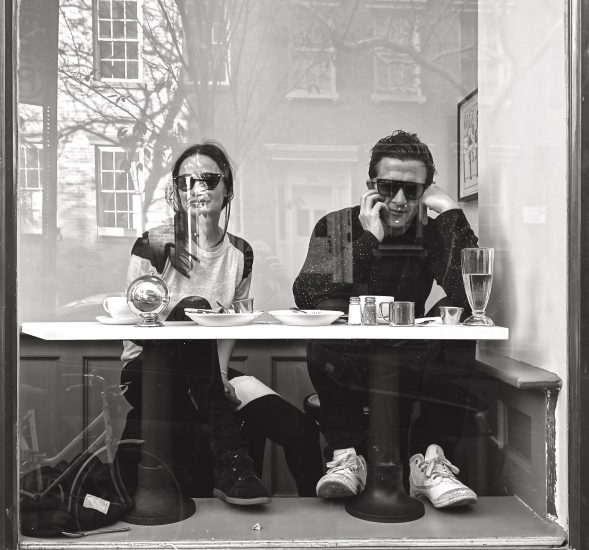A black and white photograph of a couple sitting as a table behind a large glass window.