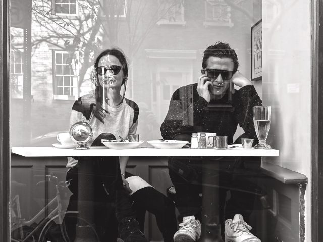 A black and white photograph of a couple sitting as a table behind a large glass window.