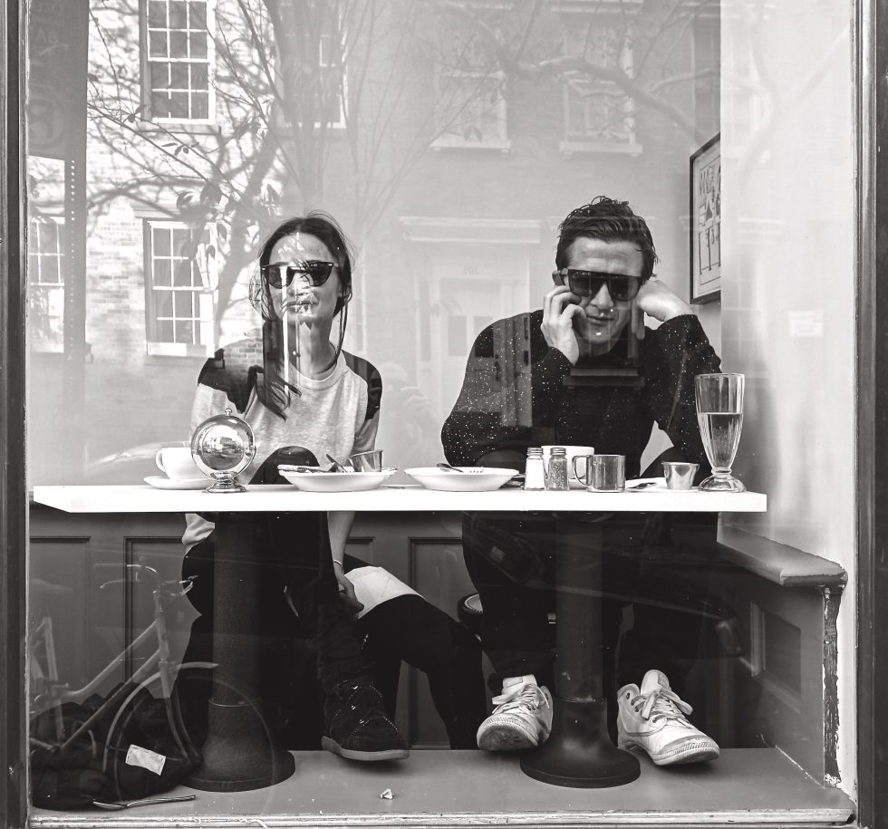 A black and white photograph of a couple sitting as a table behind a large glass window.