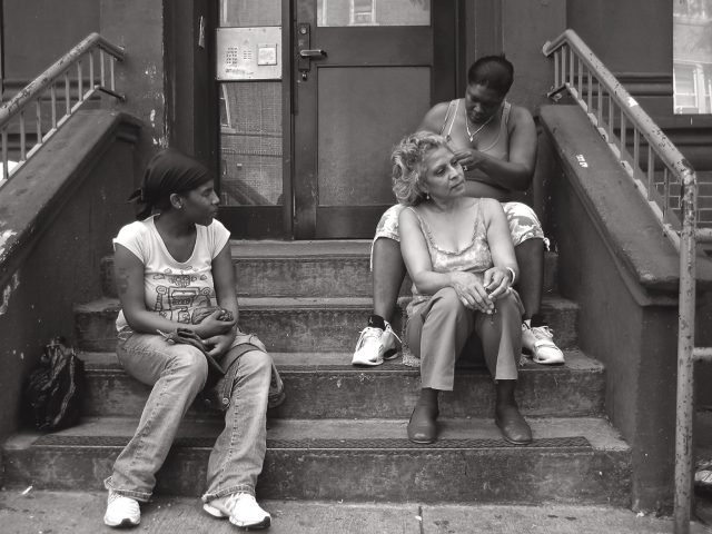 A black and white photograph of an older woman getting her hair braided on a stoop.