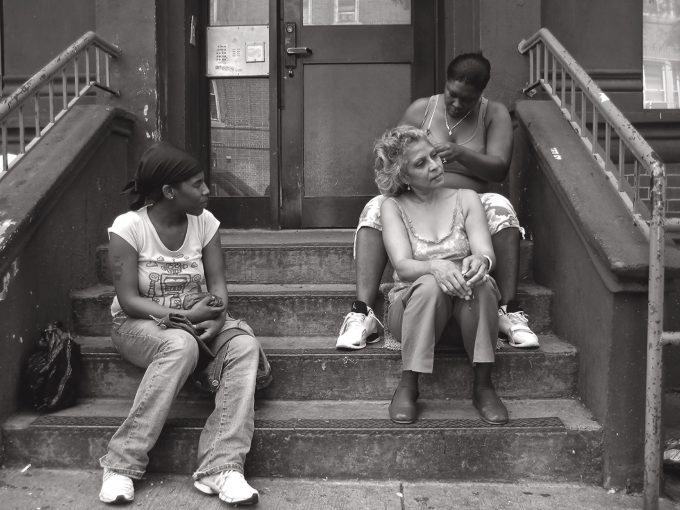A black and white photograph of an older woman getting her hair braided on a stoop.