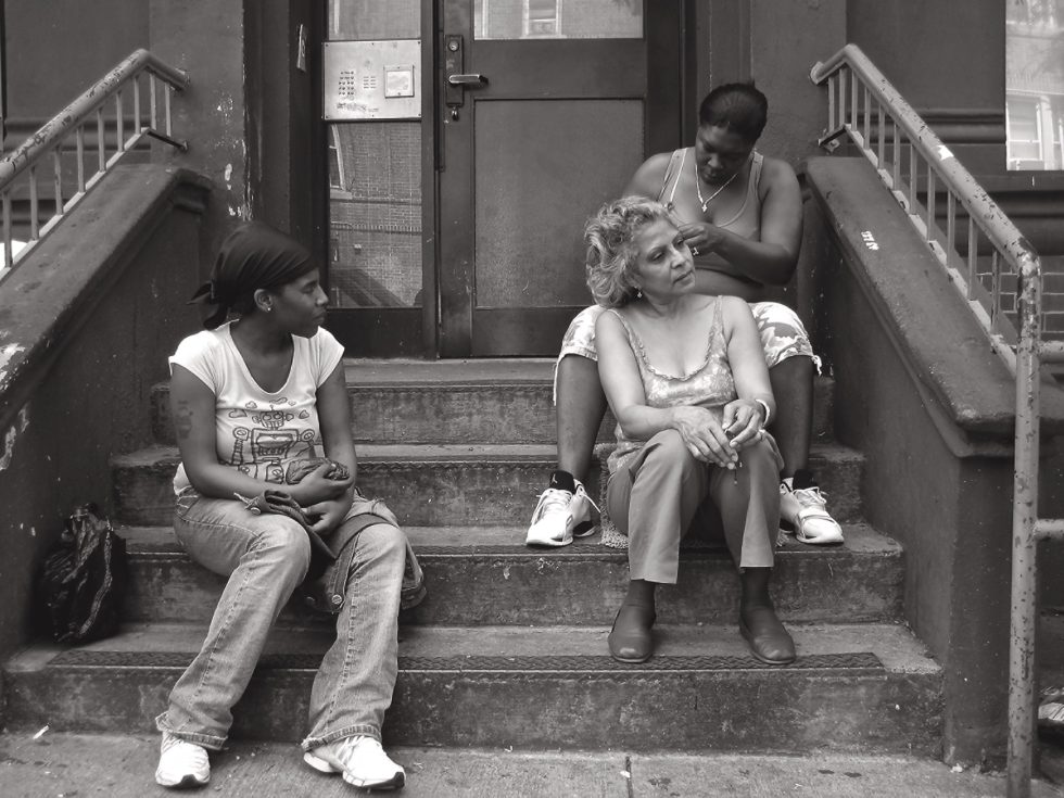 A black and white photograph of an older woman getting her hair braided on a stoop.
