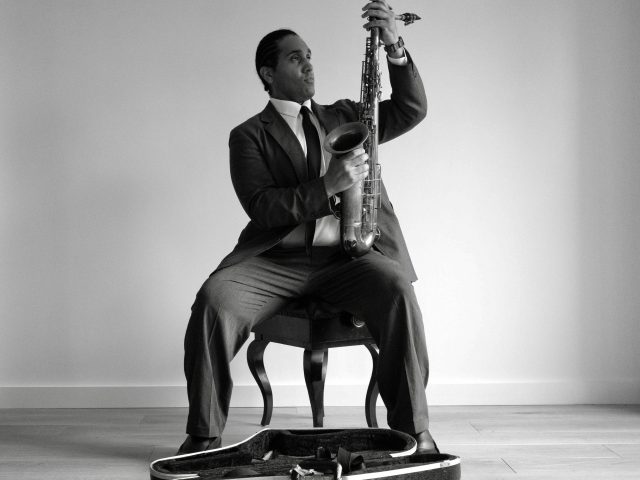 A black and white photograph of a man in a suit setting up his saxophone.