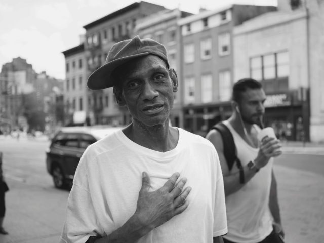 A black and white photograph of a man in a white shirt and baseball hat on a city street.