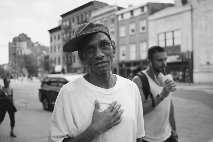 A black and white photograph of a man in a white shirt and baseball hat on a city street.