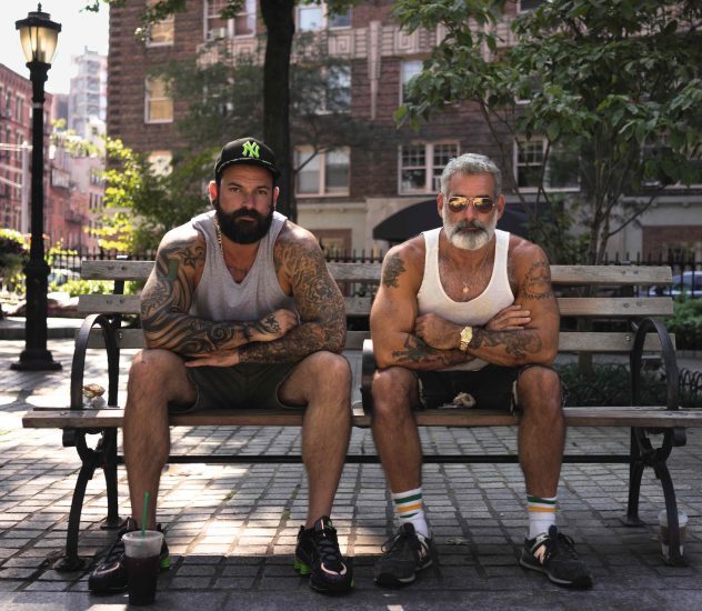A color photograph of two masculine gay men sitting on a park bench.