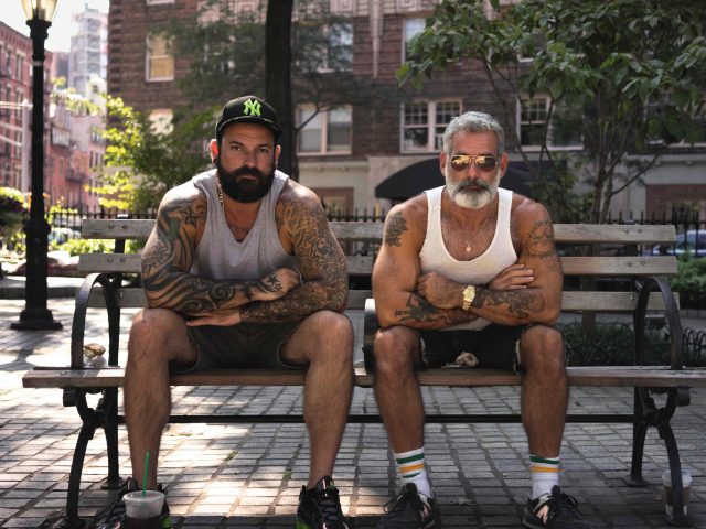 A color photograph of two masculine gay men sitting on a park bench.