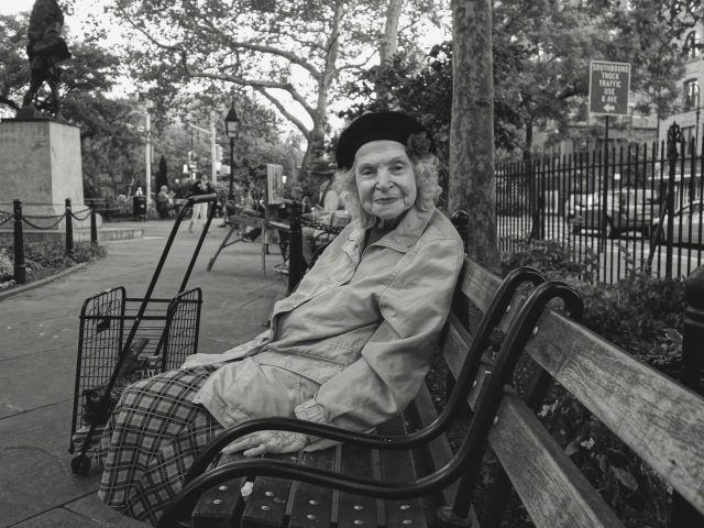 A black and white photograph of a sweet old lady sitting on a park bench smiling.