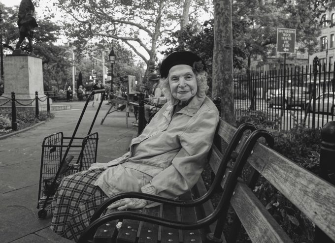 A black and white photograph of a sweet old lady sitting on a park bench smiling.