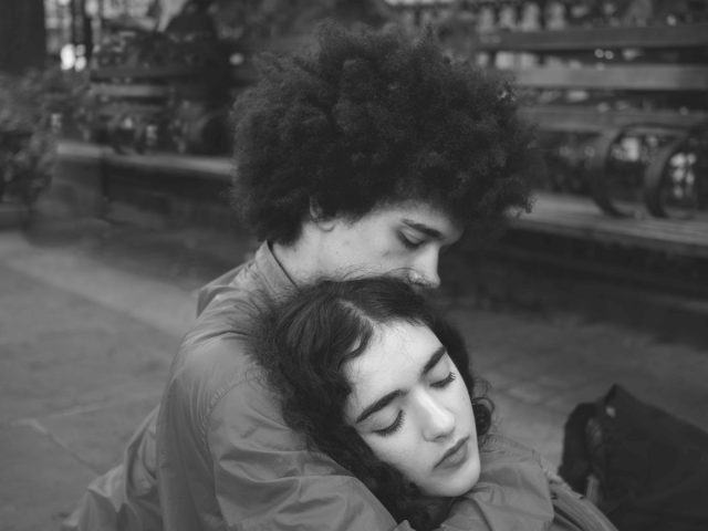 In this black and white photo, a young boy cradles the head of a girl. They are sitting in the city.