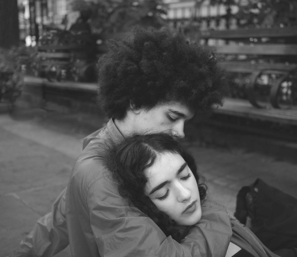 In this black and white photo, a young boy cradles the head of a girl. They are sitting in the city.