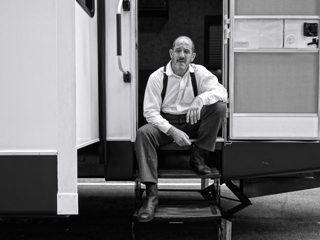 A black and white photograph of an older male actor waiting at the entrance of his trailer.