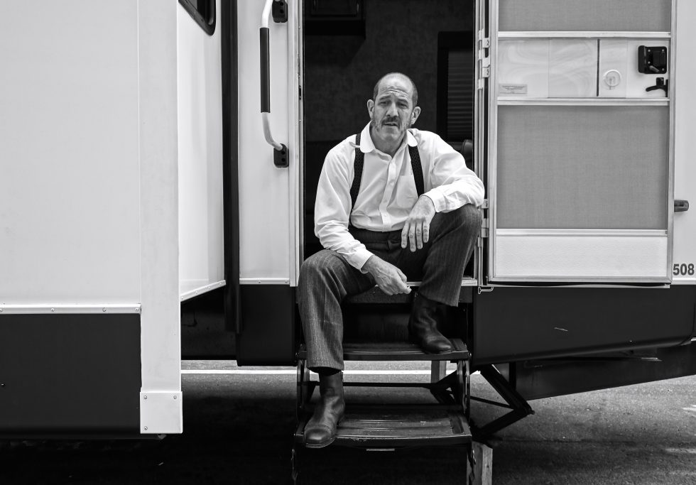 A black and white photograph of an older male actor waiting at the entrance of his trailer.