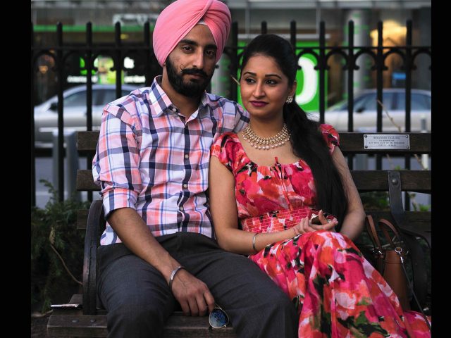 A photograph of a young couple sitting on a bench in a city park.