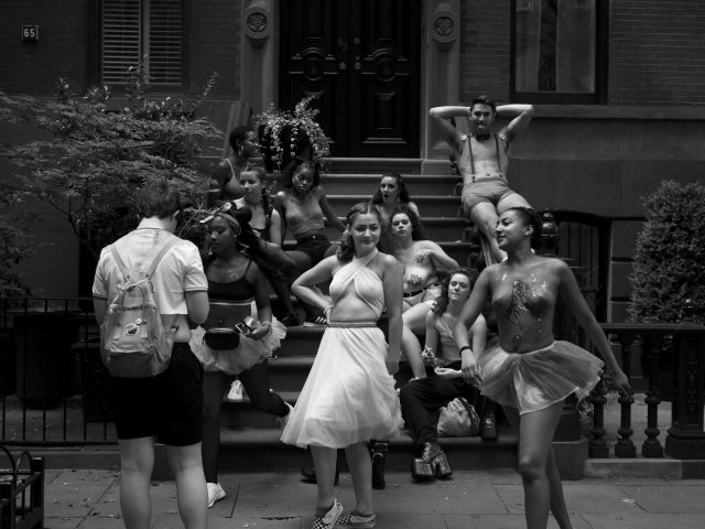 A black and white photograph of a girl in a white dress standing in front of a bunch of people on a stoop.