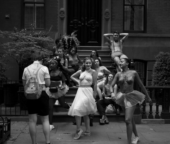 A black and white photograph of a girl in a white dress standing in front of a bunch of people on a stoop.