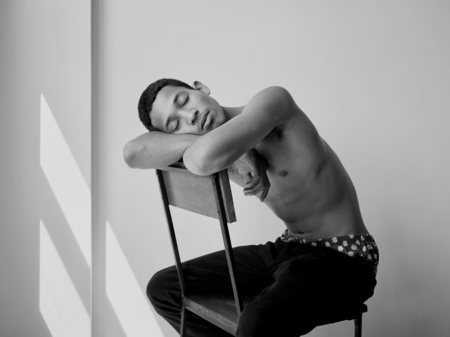 A black and white photograph of a young man sitting and resting his head on the back of a chair.