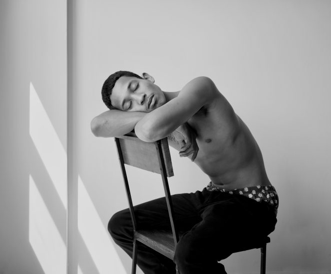 A black and white photograph of a young man sitting and resting his head on the back of a chair.