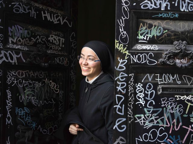 A photograph of a smiling young nun opening a grafitti-covered door to a convent.