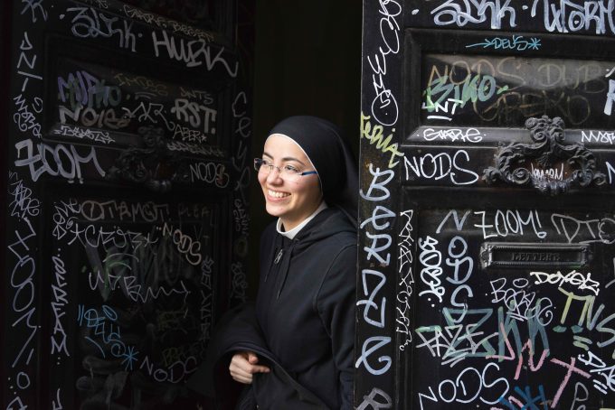 A photograph of a smiling young nun opening a grafitti-covered door to a convent.