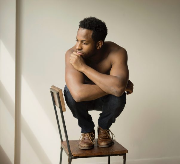 A photograph of a young man squatting in the seat of a chair.