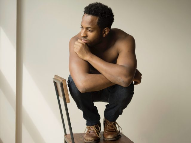 A photograph of a young man squatting in the seat of a chair.