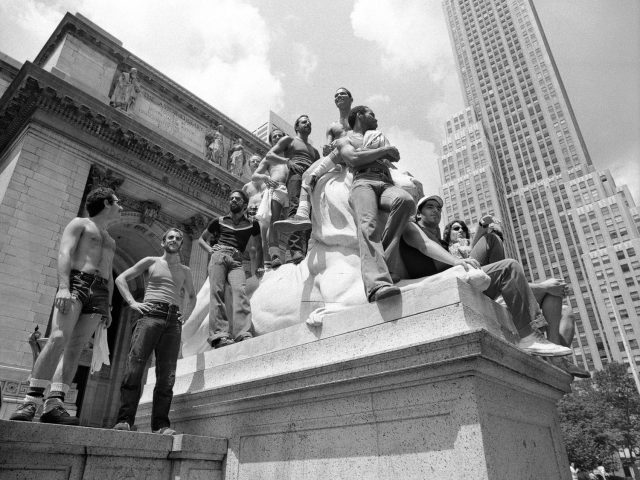A black and white photograph of a group of queer men watching a pride parade from the steps of a library.