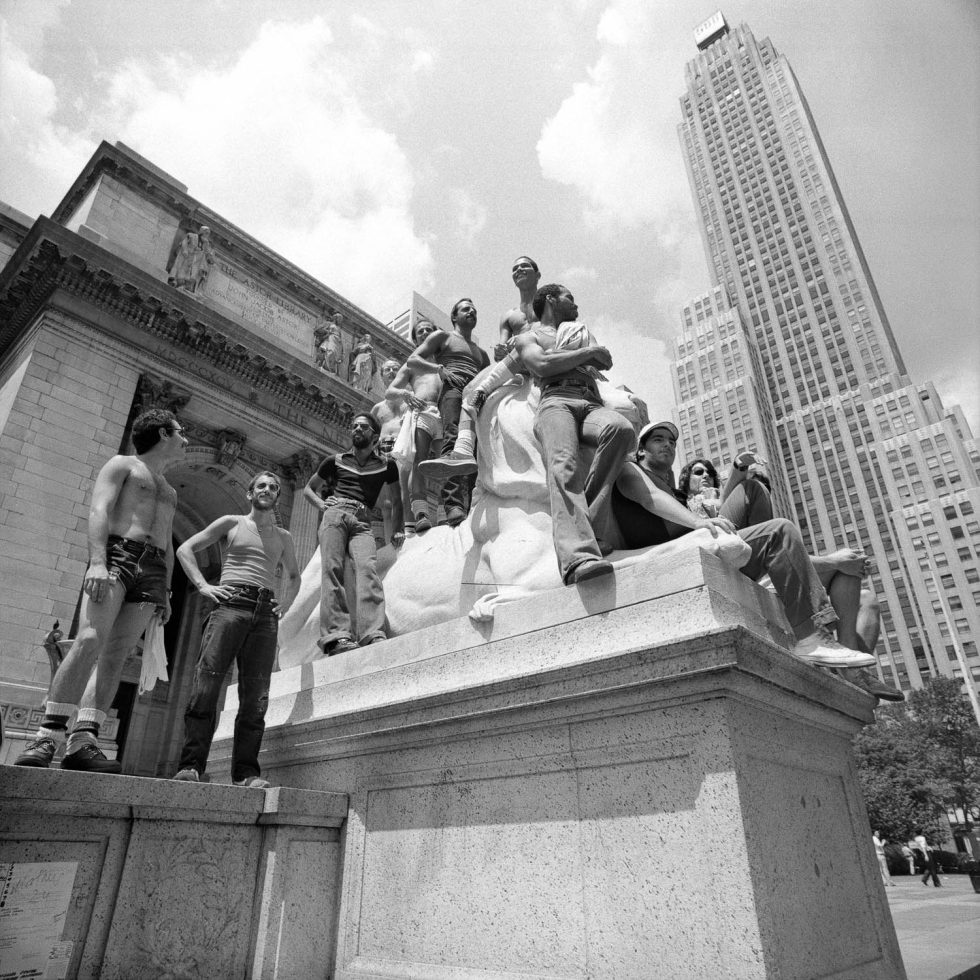 A black and white photograph of a group of queer men watching a pride parade from the steps of a library.