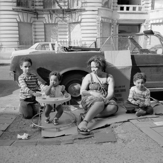 A black and white photograph of a family sitting on cardboard next to a car.