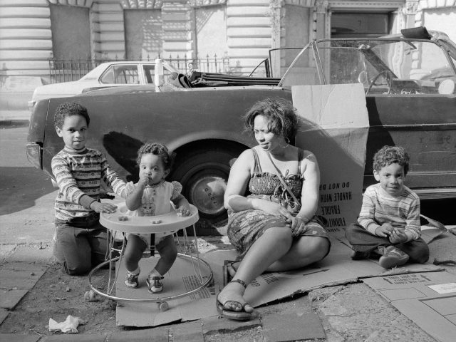 A black and white photograph of a family sitting on cardboard next to a car.