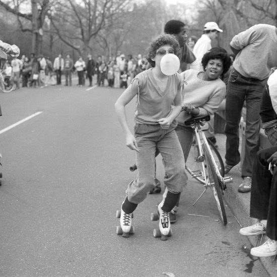 A black and white photograph of a boy blowing a bubble gum bubble in roller skates.