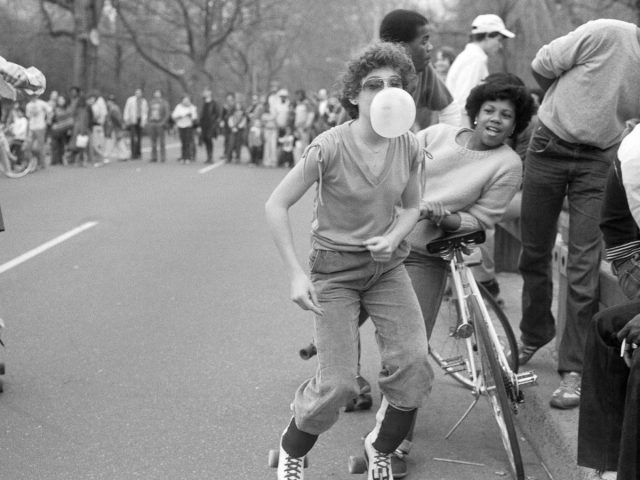 A black and white photograph of a boy blowing a bubble gum bubble in roller skates.