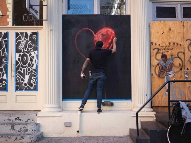 A photograph of a man spray painting a red heart onto a boarded up storefront.