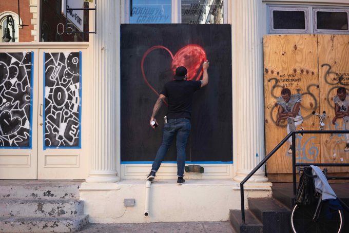 A photograph of a man spray painting a red heart onto a boarded up storefront.