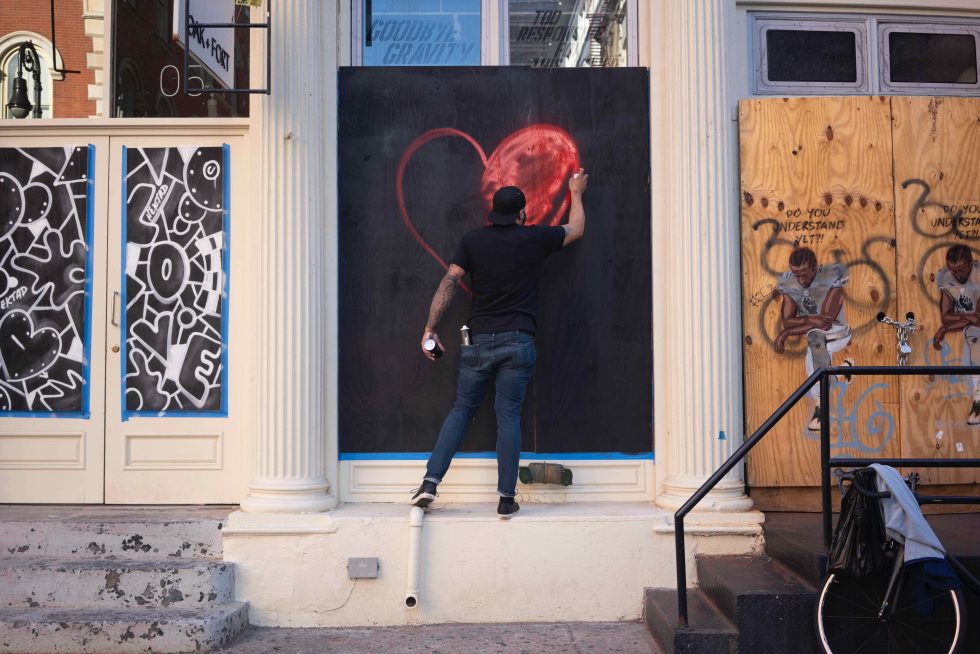 A photograph of a man spray painting a red heart onto a boarded up storefront.