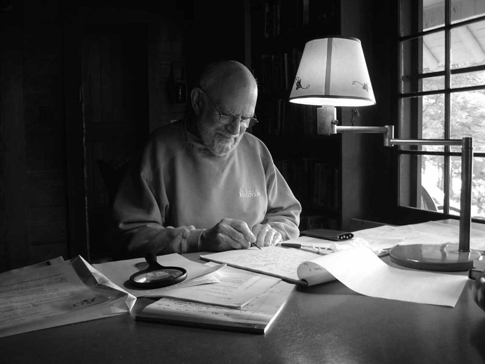 A black and white photograph of an older man sitting at a desk next to a lamp.