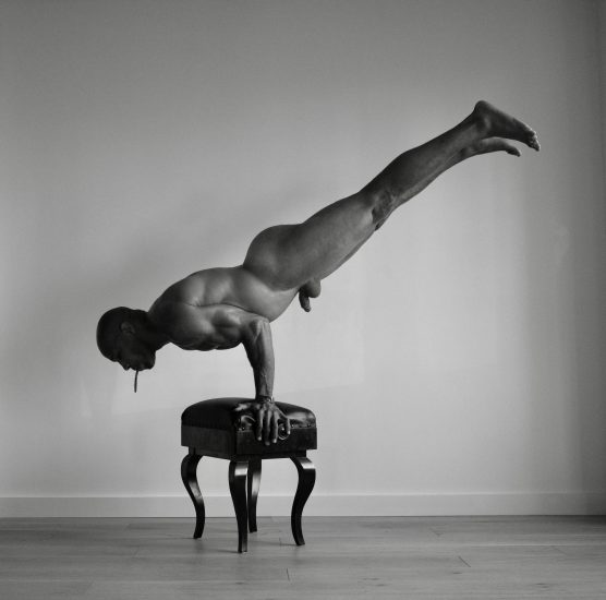 A black and white photograph of a nude man doing a handstand on a piano bench.