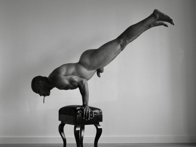 A black and white photograph of a nude man doing a handstand on a piano bench.