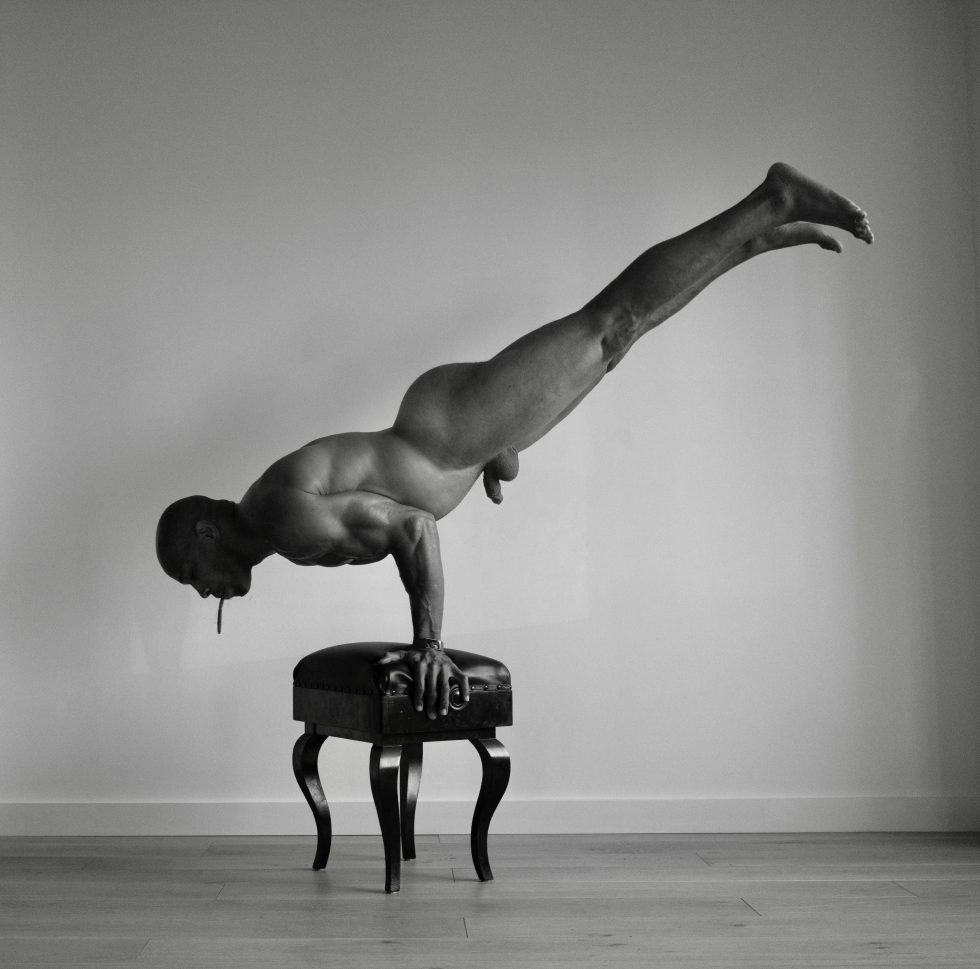 A black and white photograph of a nude man doing a handstand on a piano bench.