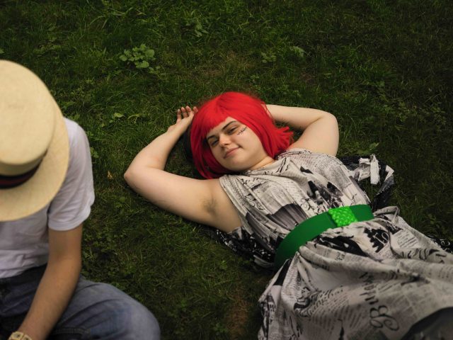 A colorful photograph of a young woman with red hair and a green belt lying in grass.