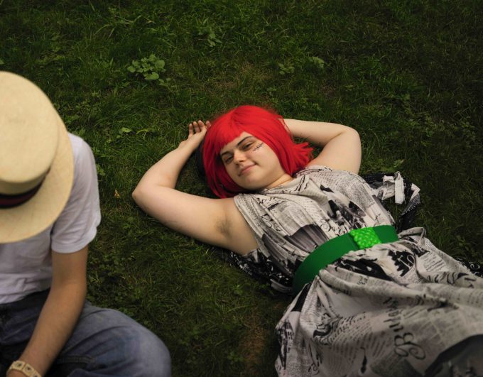 A colorful photograph of a young woman with red hair and a green belt lying in grass.
