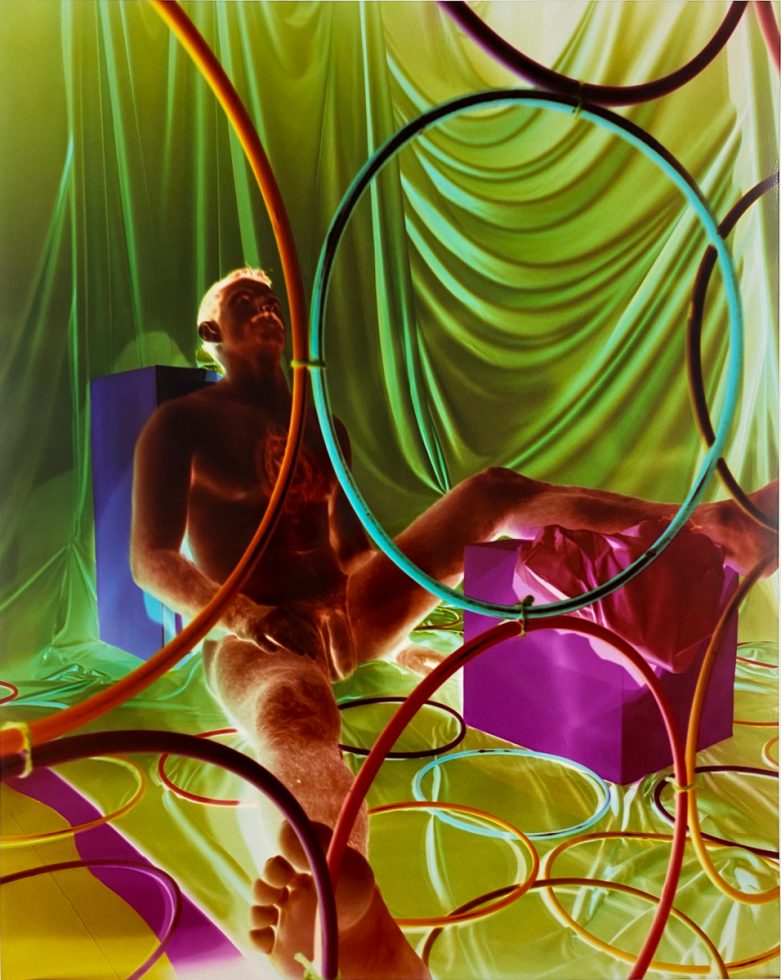 A colorful image of a nude man sitting on the floor with circles creating a design on top of the image.