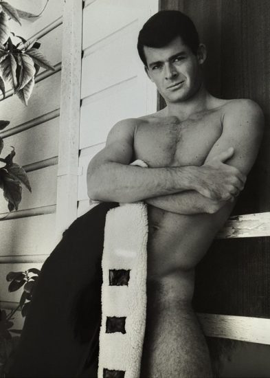 A black and white photograph of a nude man, covering his lower body with a jacket.