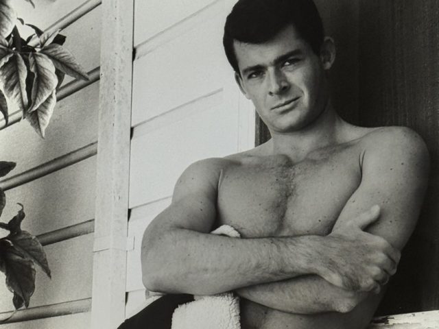 A black and white photograph of a nude man, covering his lower body with a jacket.